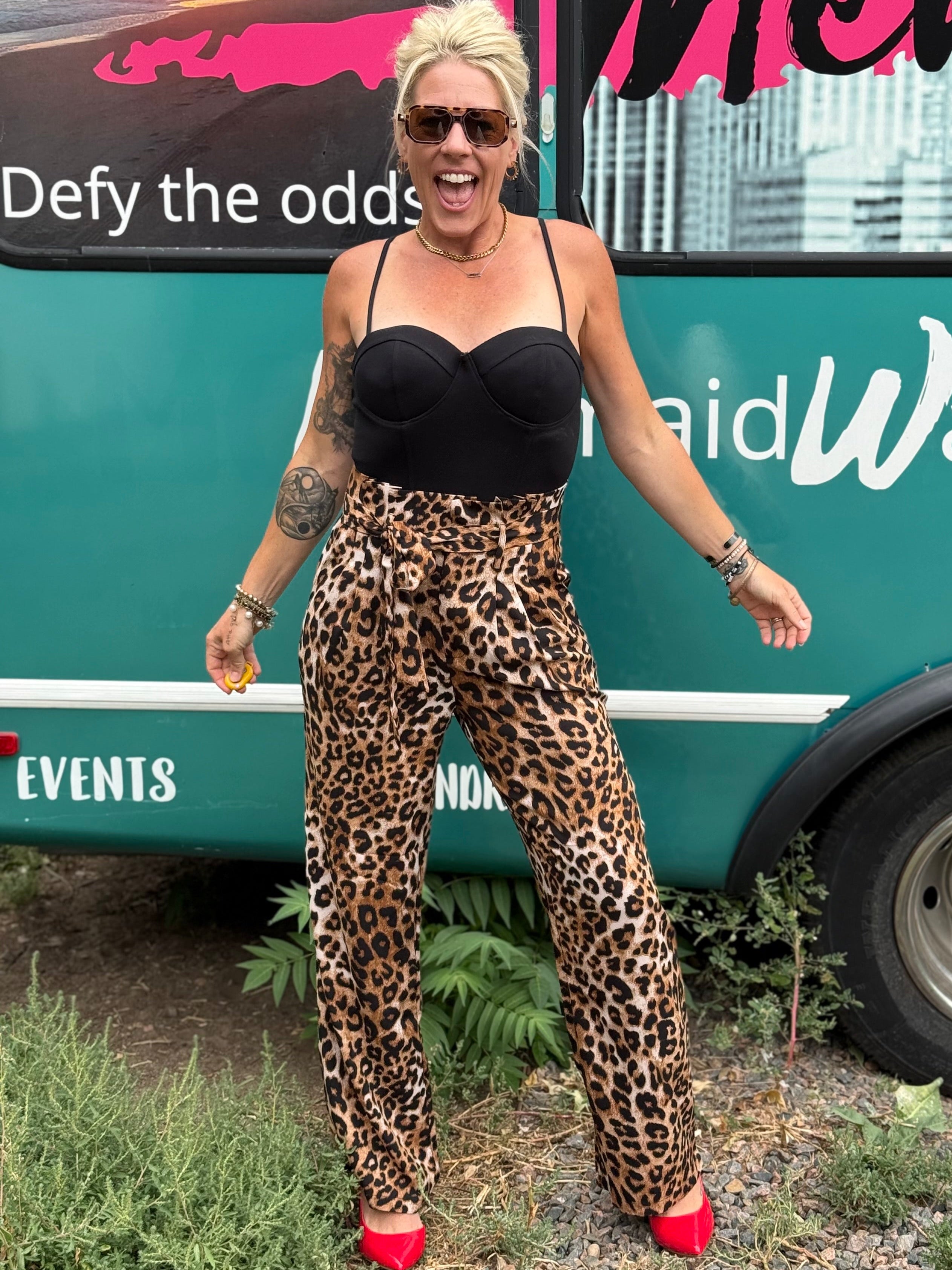 Woman in leopard print pants and black top standing in front of a green bus with 'Mermaid Walk' branding.