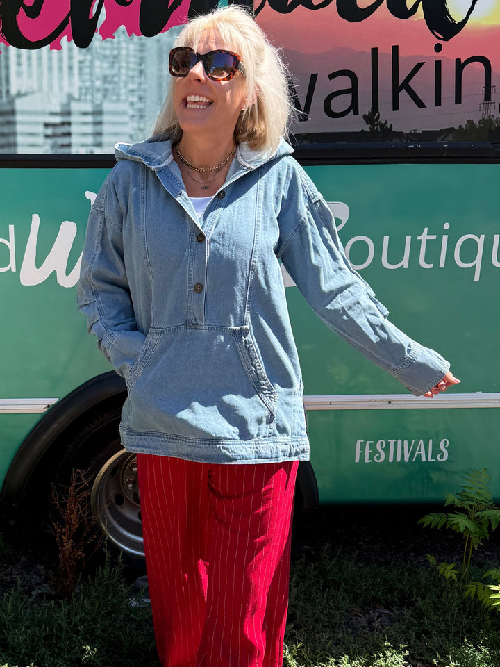 A woman wearing a light blue denim jacket and bright red pants stands in front of a green bus with &