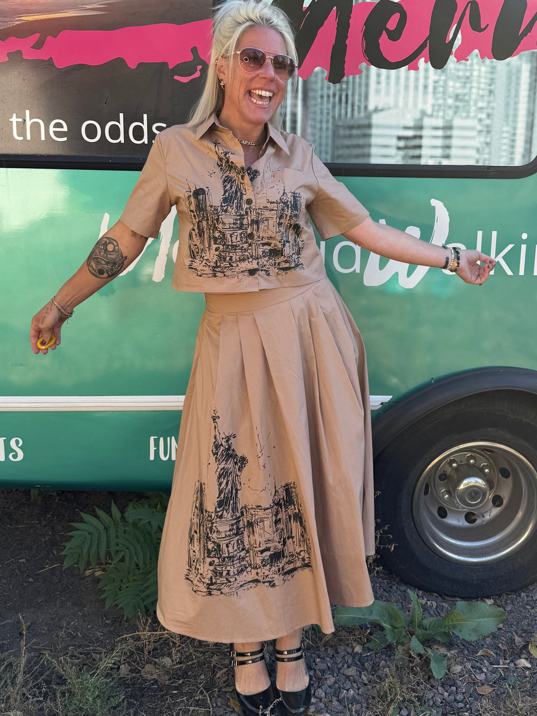 Woman in a beige dress standing next to a green bus with text on the side.