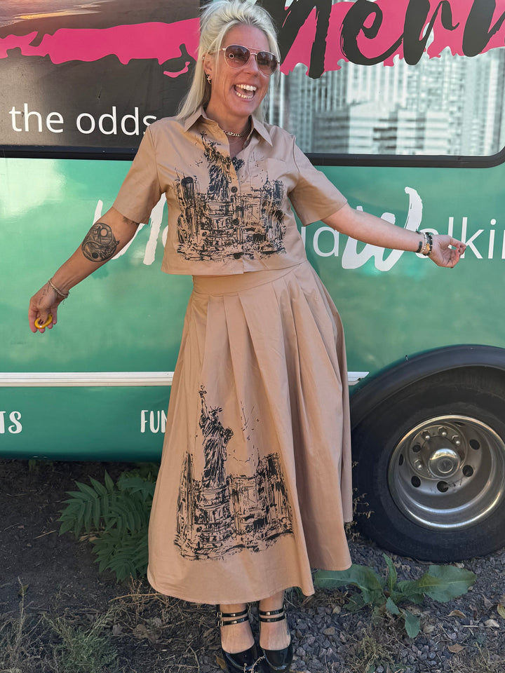 Woman in a beige dress standing next to a green bus with text on the side.