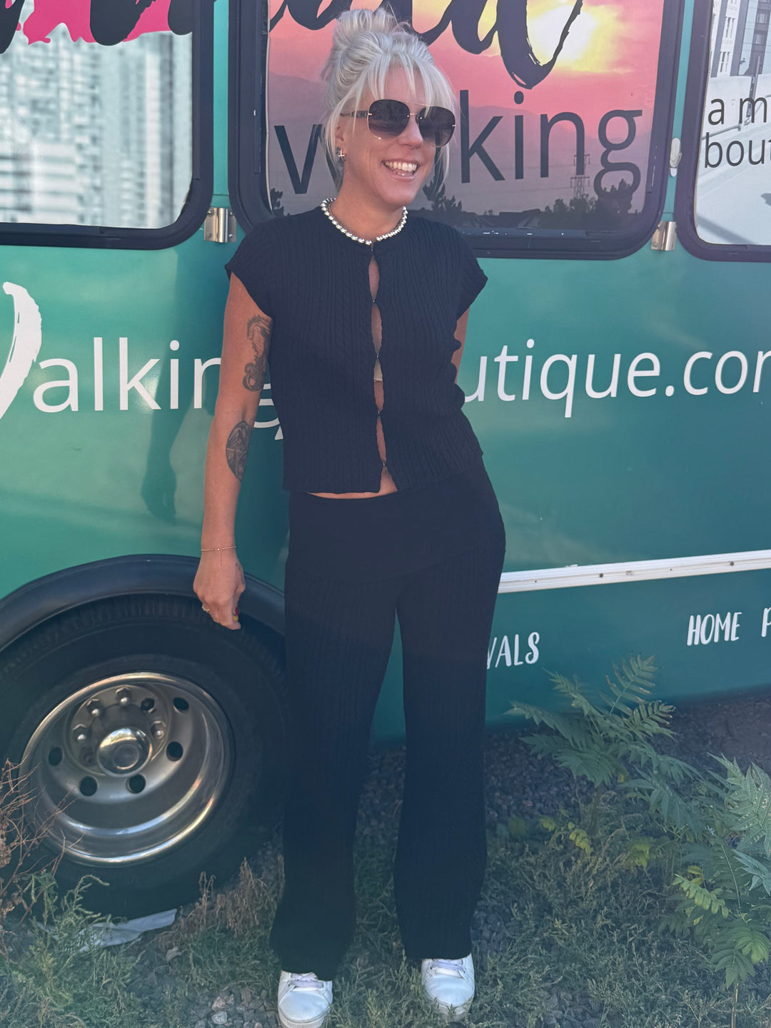 A woman wearing a black two-piece outfit with white sneakers stands in front of a teal mobile boutique bus. The bus has large windows with a sunset reflection and text including 'Mermaid Walking Boutique' and 'mermaidwalkingboutique.