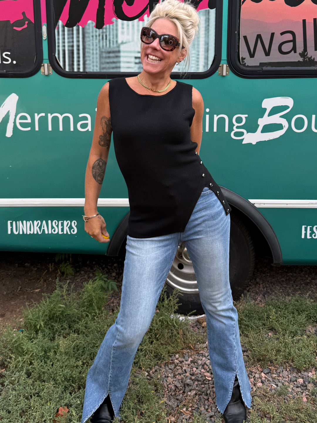 A woman with short blonde hair and sunglasses poses in front of a teal event bus with &