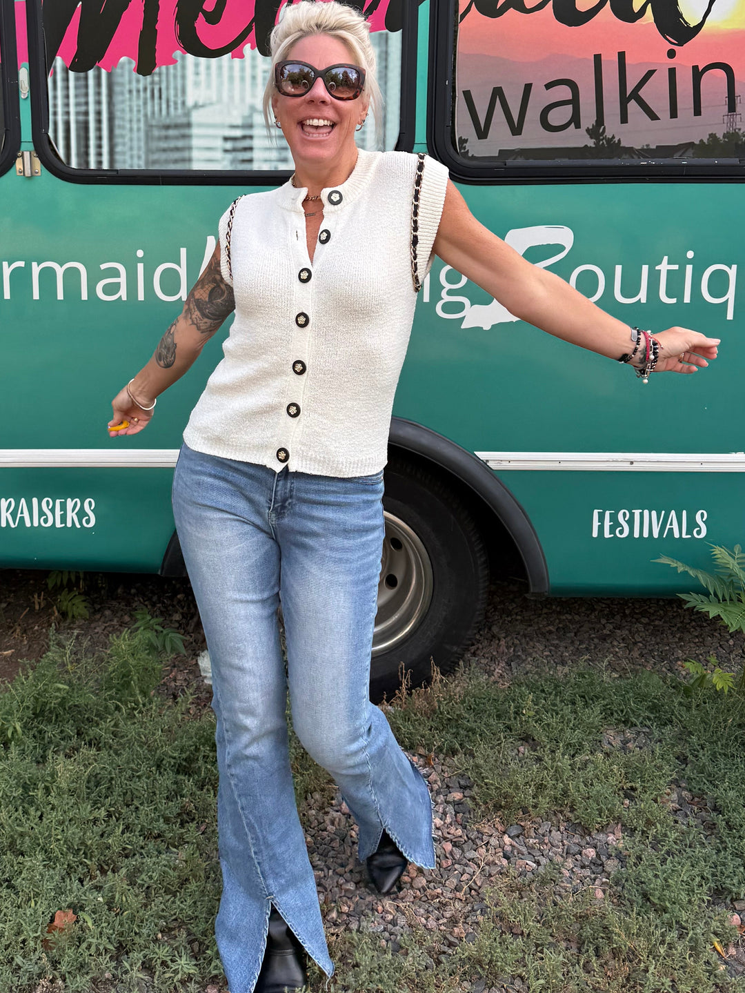 A woman posing in front of a teal bus with 'Mermaid Walking Boutique' printed on the side, wearing a cream sleeveless vest and blue flared jeans.