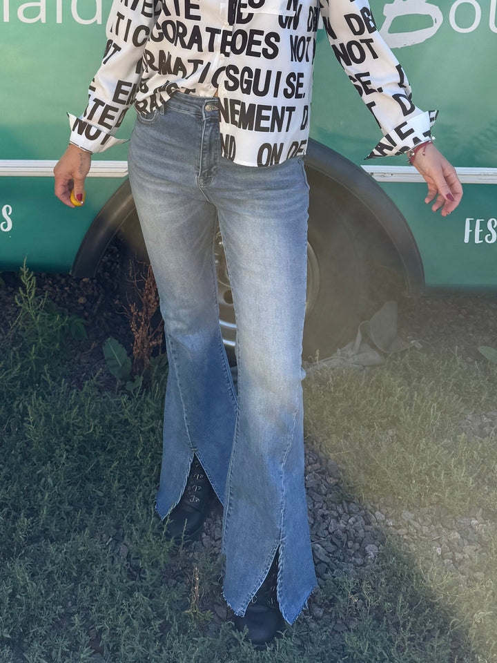 A woman wearing a white long-sleeve top with black text and light blue jeans stands in front of a teal bus with &