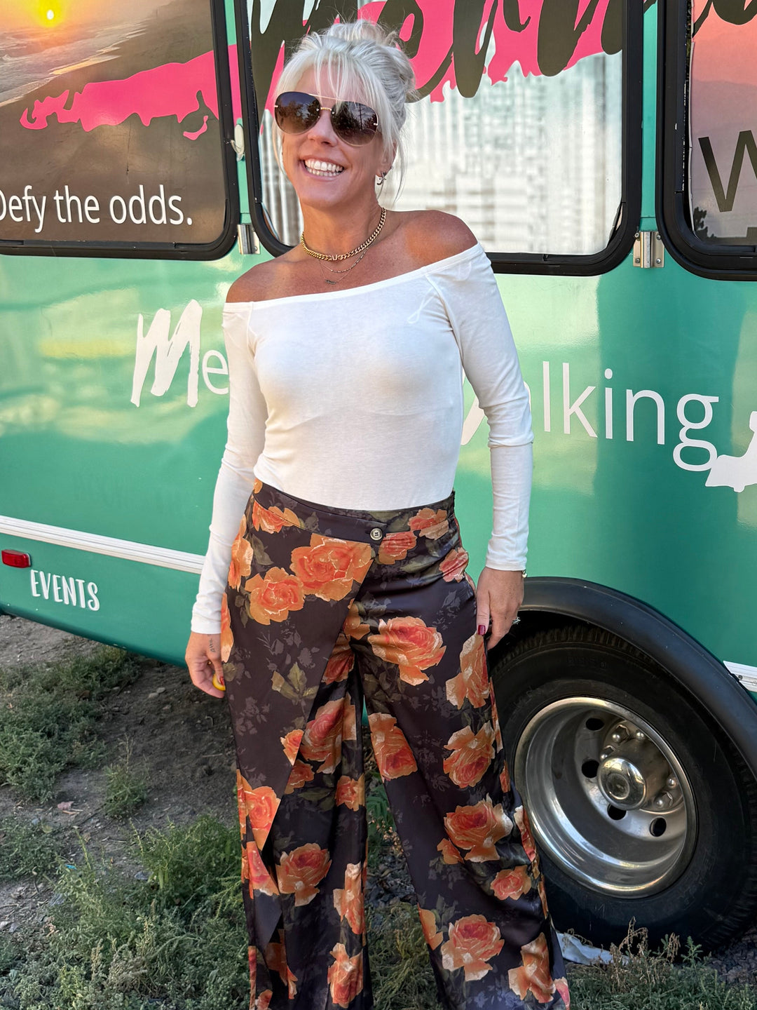 A woman stands in front of a teal bus with large windows and colorful sunset graphics. She is wearing a white off-the-shoulder top and wide-leg floral pants with brown boots.
