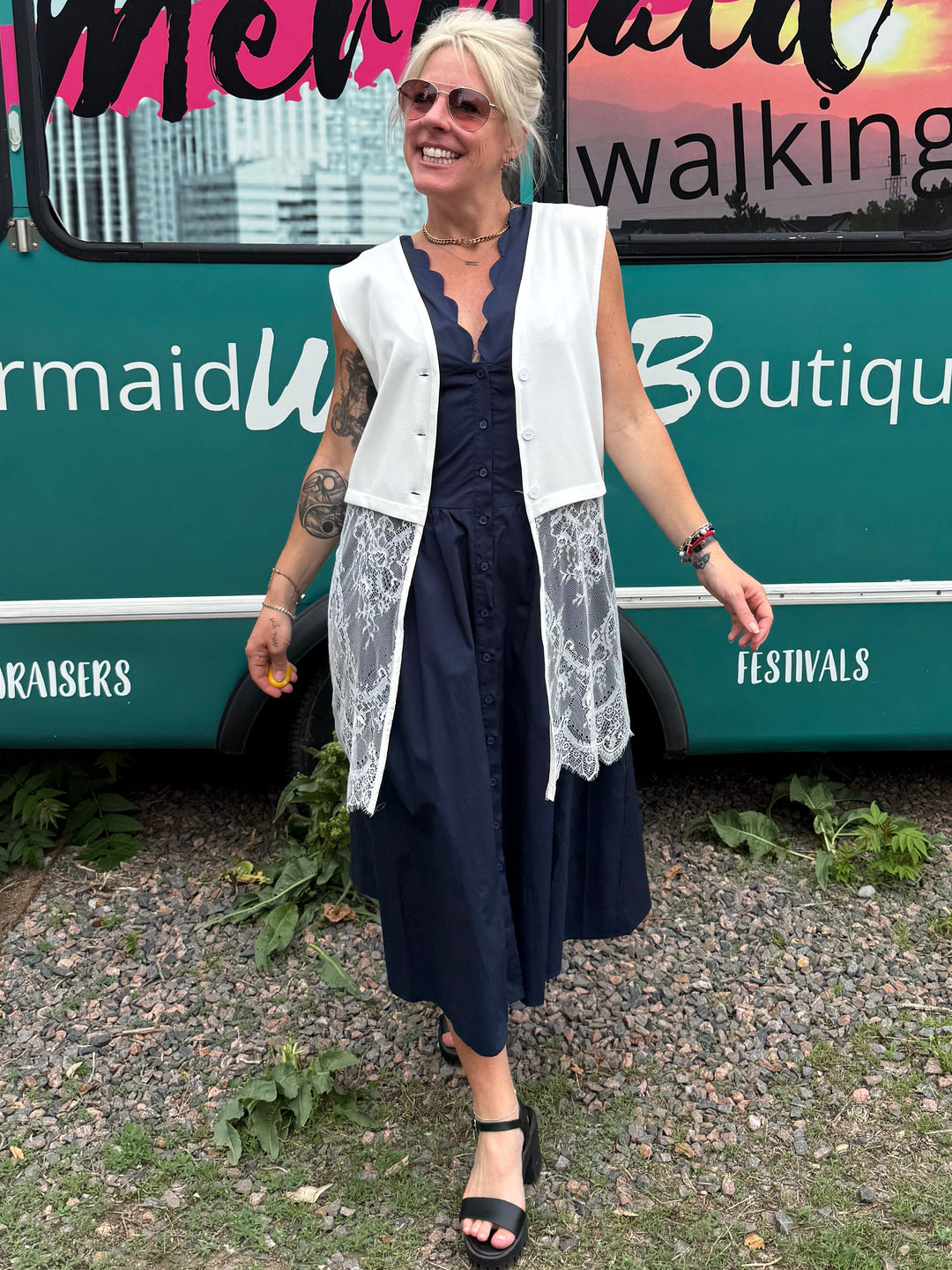 A woman stands in front of a teal bus with 'Mermaid Walking Boutique' printed on the side, wearing a navy blue midi dress with a white lace-trimmed sleeveless vest and black strappy heels.