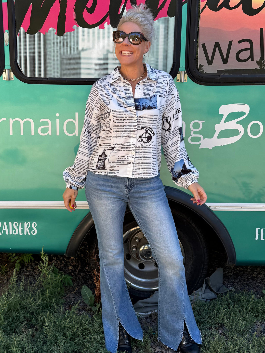 A woman wearing a patterned long-sleeve button-up shirt, blue flared jeans, and black shoes stands in front of a teal bus with 'Mermaid Walking Boutique' written on the side.