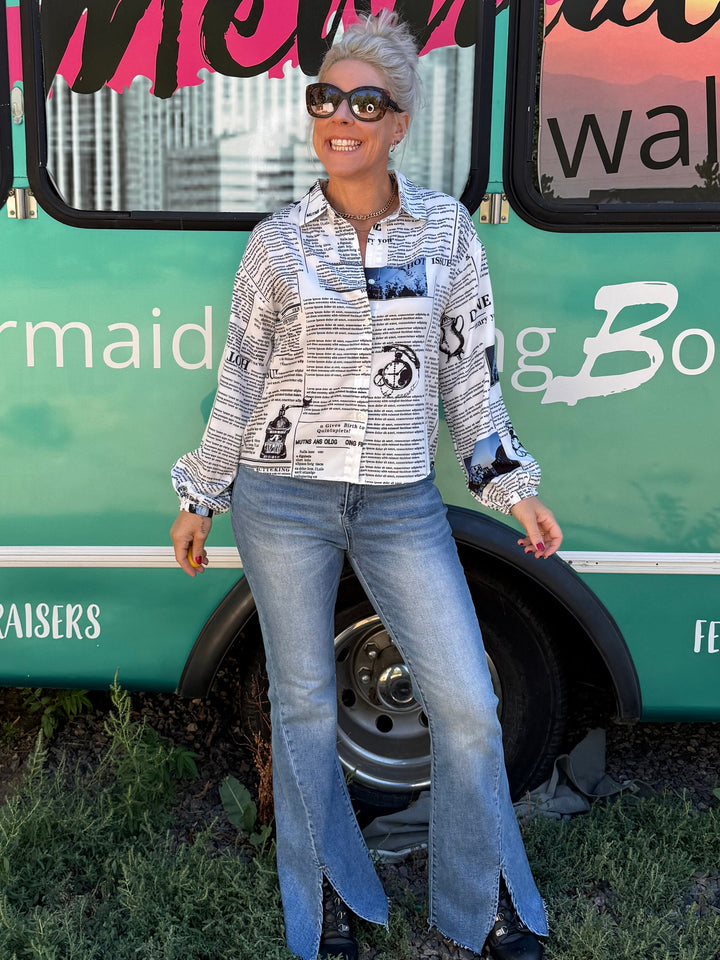 A woman wearing a patterned long-sleeve button-up shirt, blue flared jeans, and black shoes stands in front of a teal bus with &