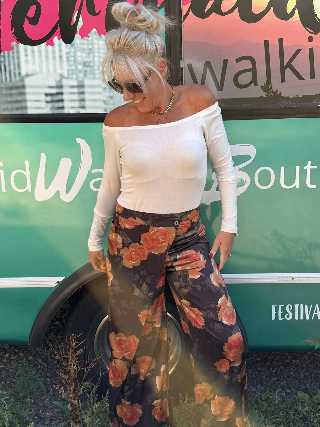 A woman stands outdoors in front of a teal bus with 'Mermaid Walking Boutique' written on it, wearing a white off-the-shoulder long-sleeve top and dark wide-leg pants with a large red and orange floral print.