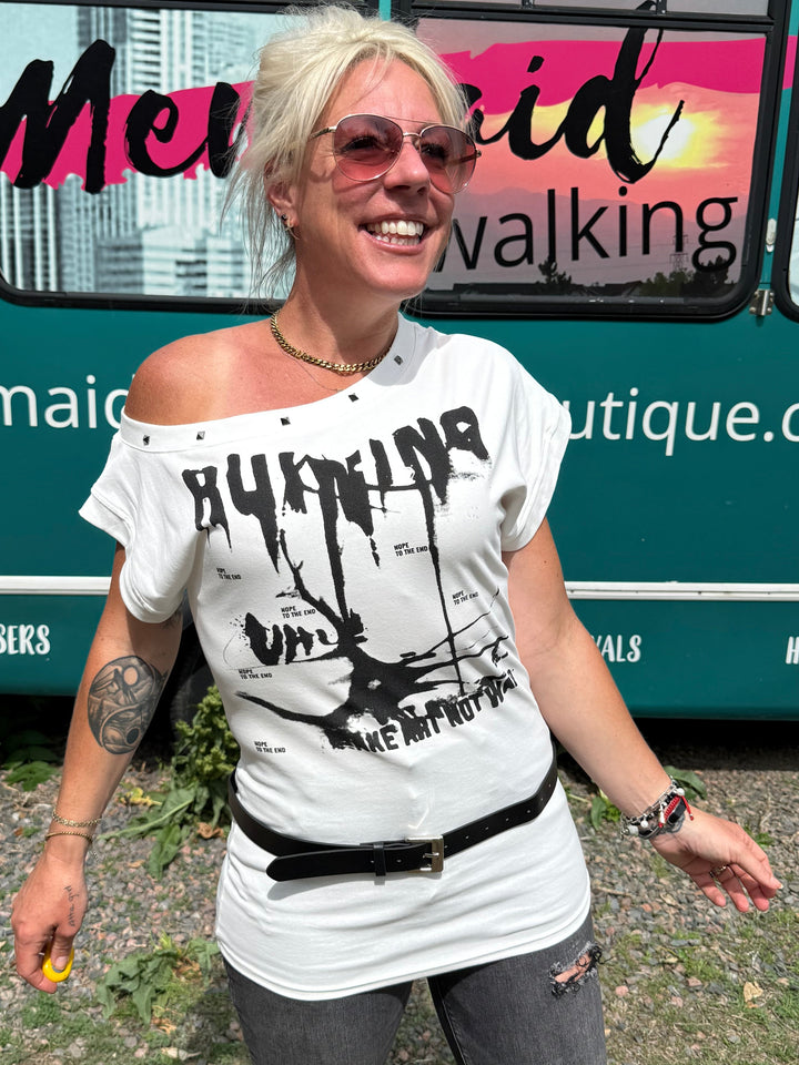 A woman stands outdoors in front of a teal bus wearing a white off-the-shoulder graphic T-shirt with black text and distressed gray jeans.