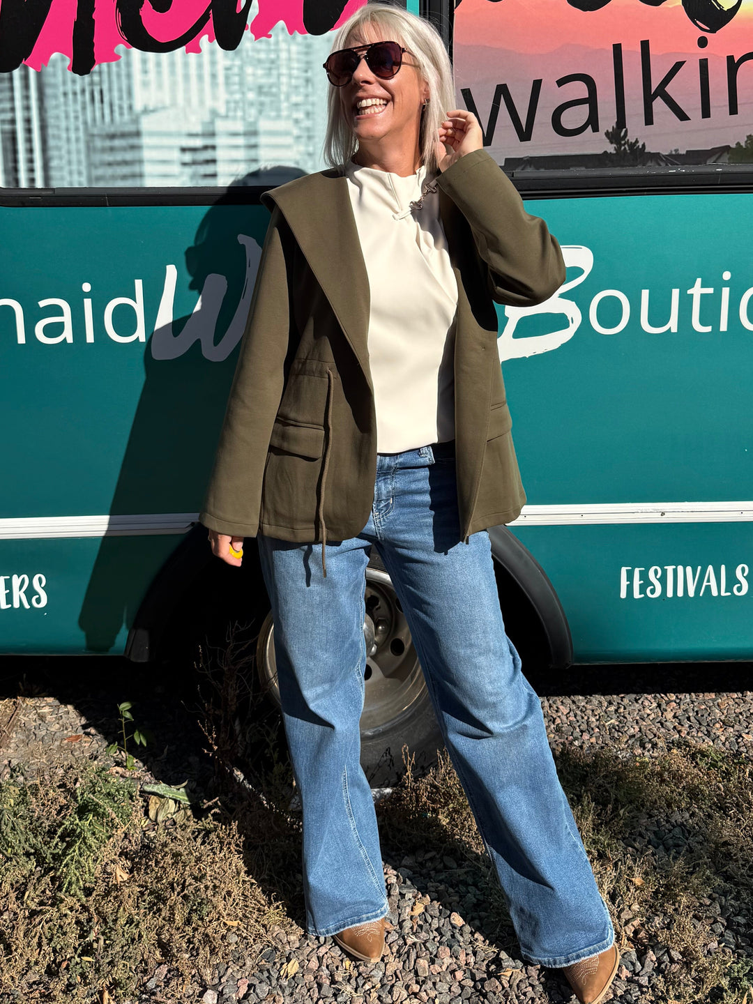 A woman wearing a green blazer, white top, and blue jeans stands in front of a teal bus with &