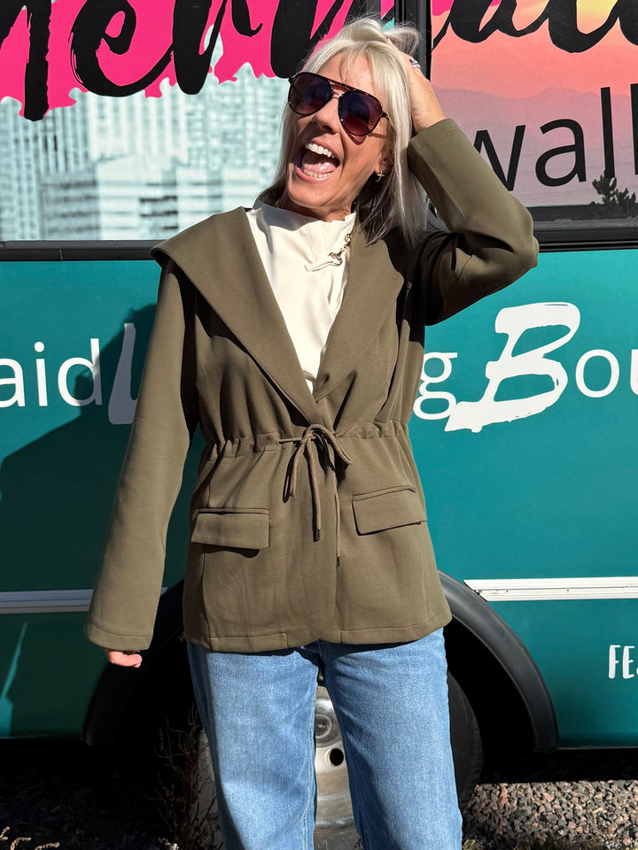 A woman wearing a green belted blazer, white top, and blue jeans stands in front of a teal bus with &