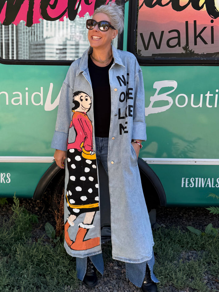 A woman wearing a long light blue denim coat over a black top and jeans stands in front of a turquoise bus with &