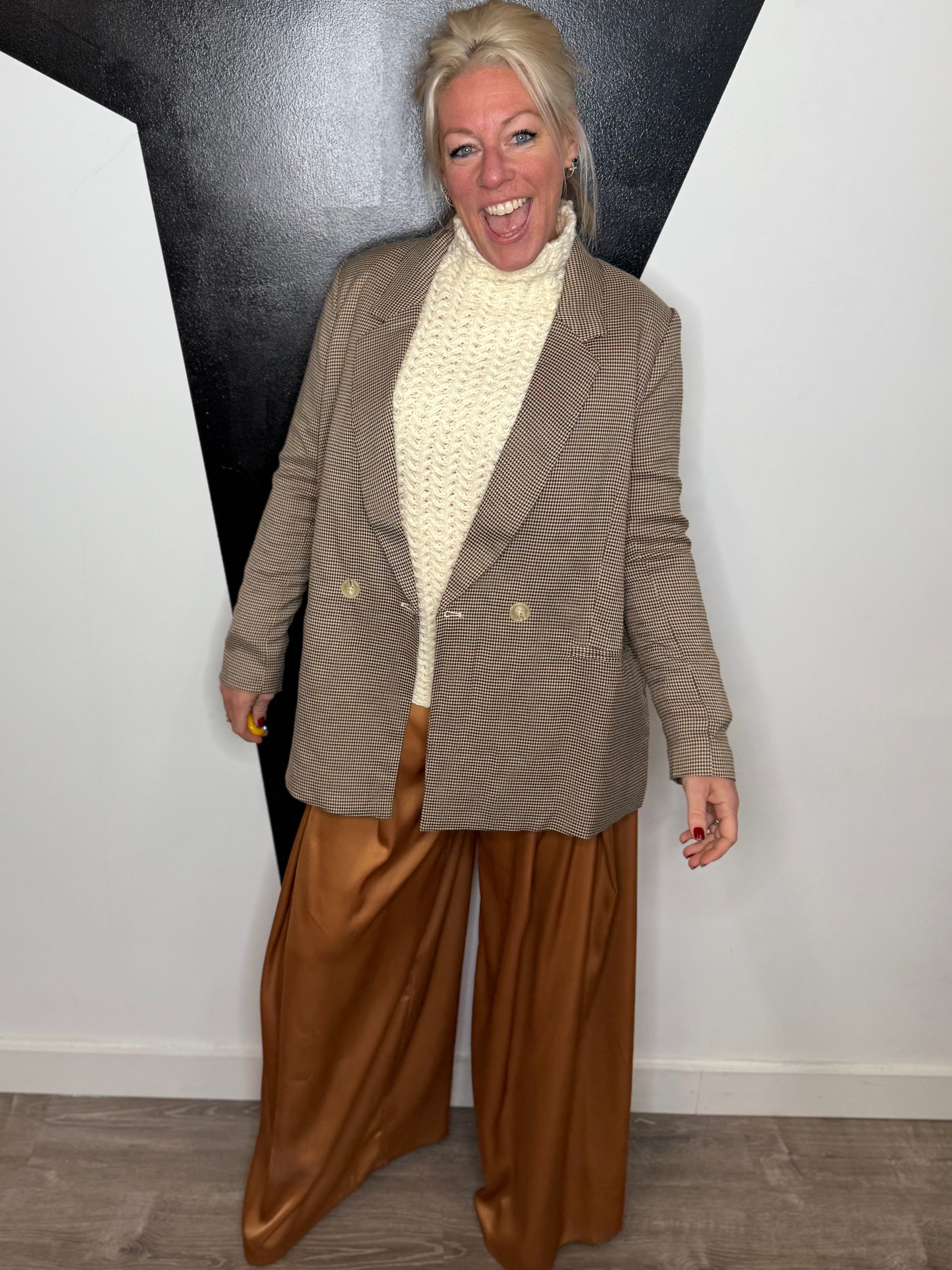 Woman in a brown blazer and gold pants standing against a black and white wall.