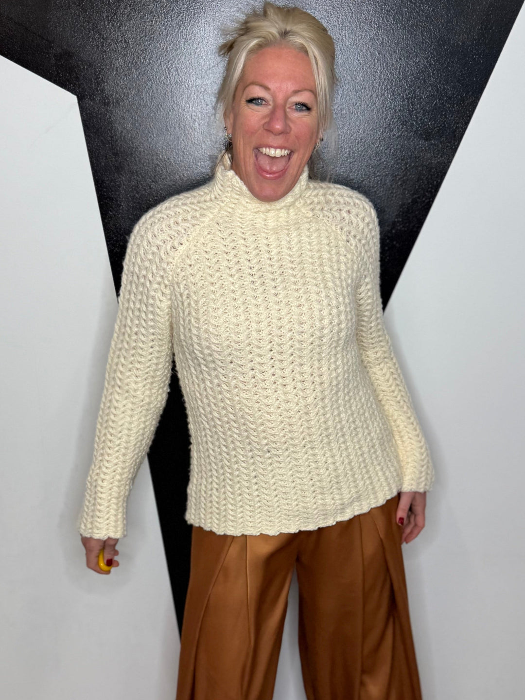 Person wearing a cream sweater and brown pants standing against a black and white geometric wall.