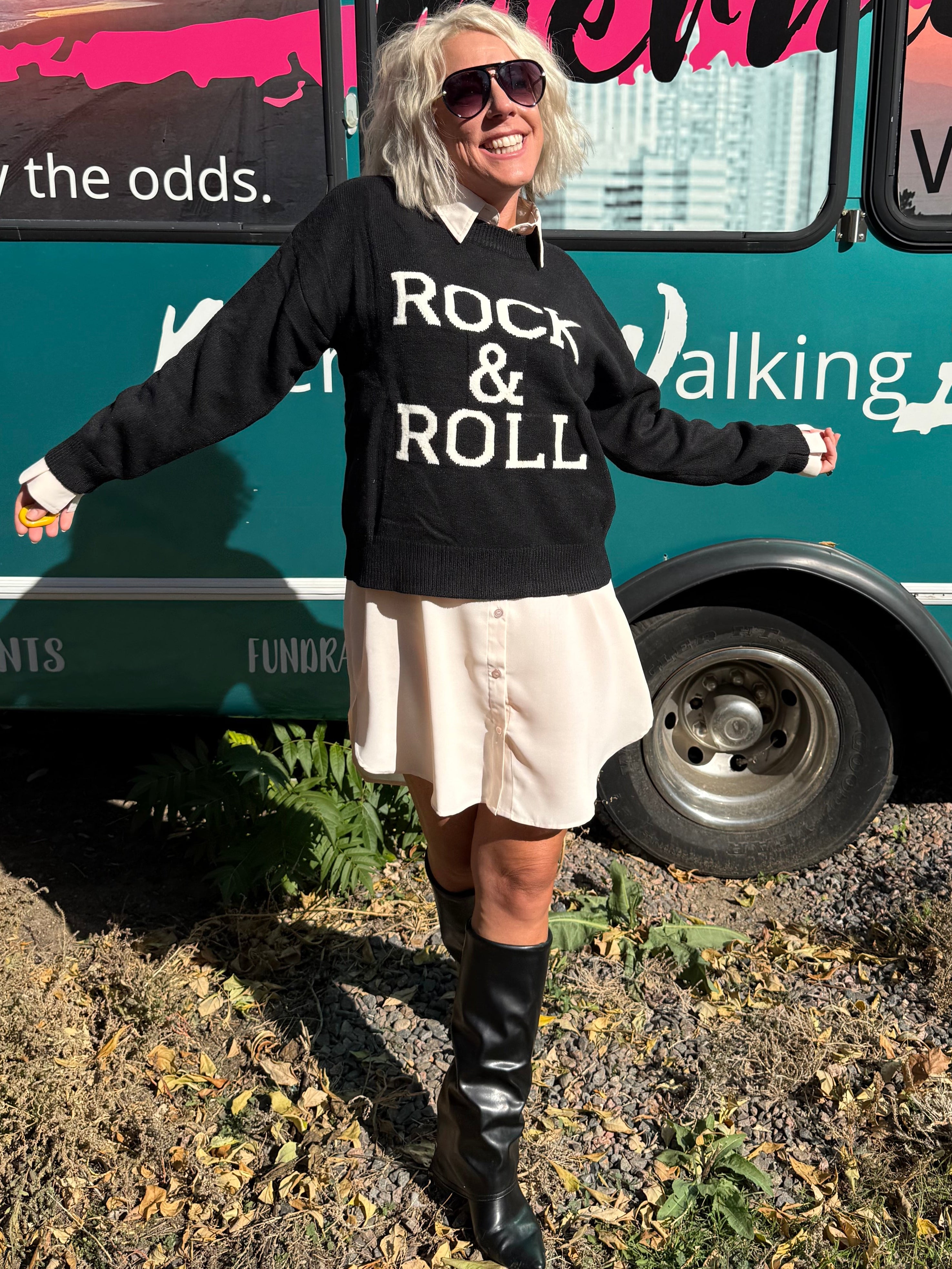 Person wearing a 'Rock & Roll' sweatshirt standing in front of a teal bus with reflective surfaces.
