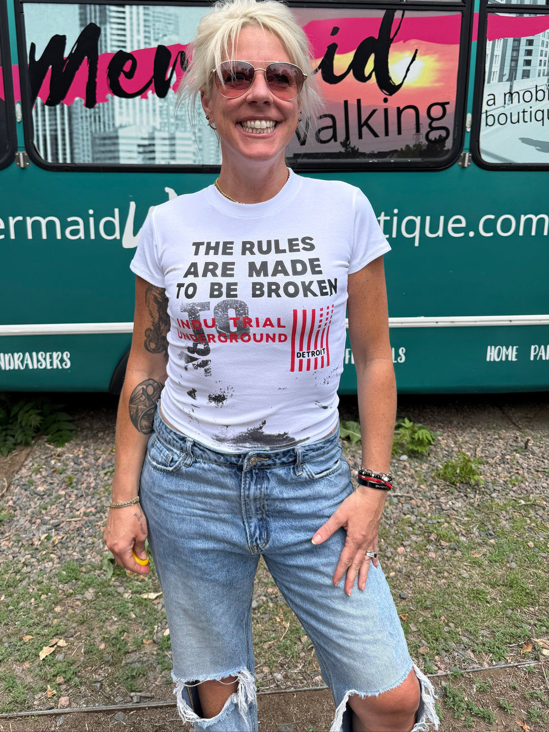 A woman with short blonde hair and sunglasses stands in front of a teal bus, wearing a white graphic t-shirt with distressed black text and ripped light-wash denim jeans.