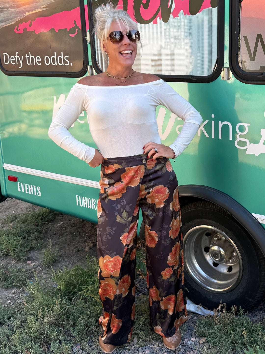 A woman stands in front of a teal bus with large windows and colorful sunset graphics. She is wearing a white off-the-shoulder long-sleeve top and wide-leg patterned pants with orange, brown, and black tones.