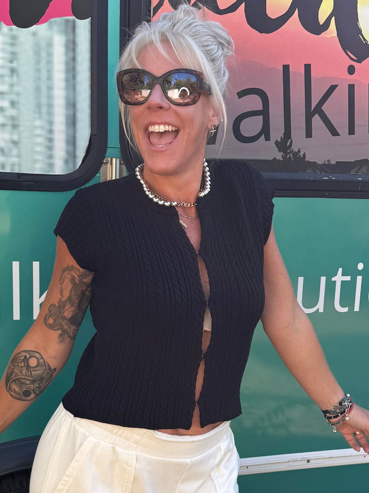 A woman with short gray hair and sunglasses stands in front of a teal bus with &