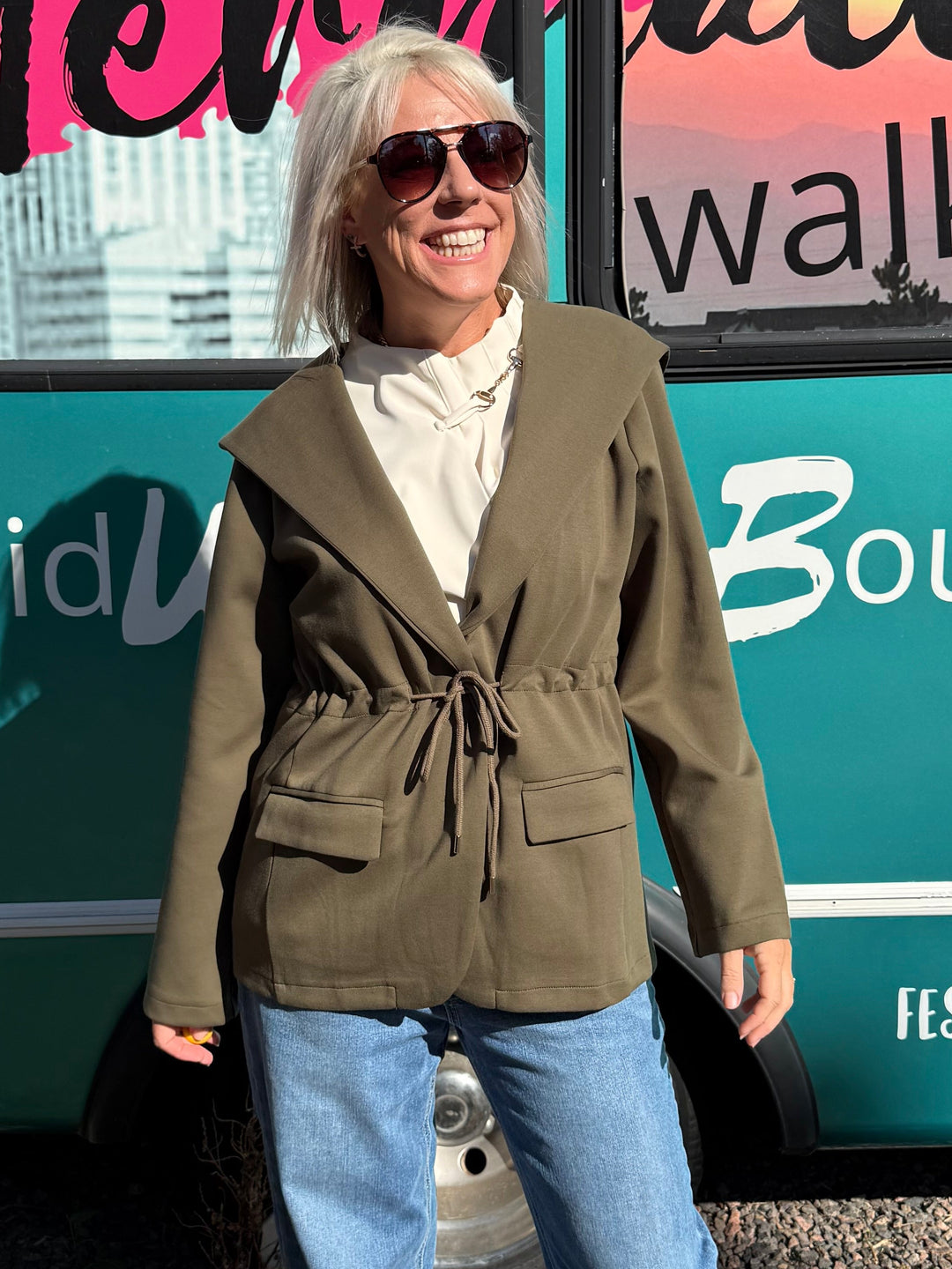 A woman wearing an olive green wrap-style blazer, cream shirt, and blue jeans stands in front of a teal bus with &