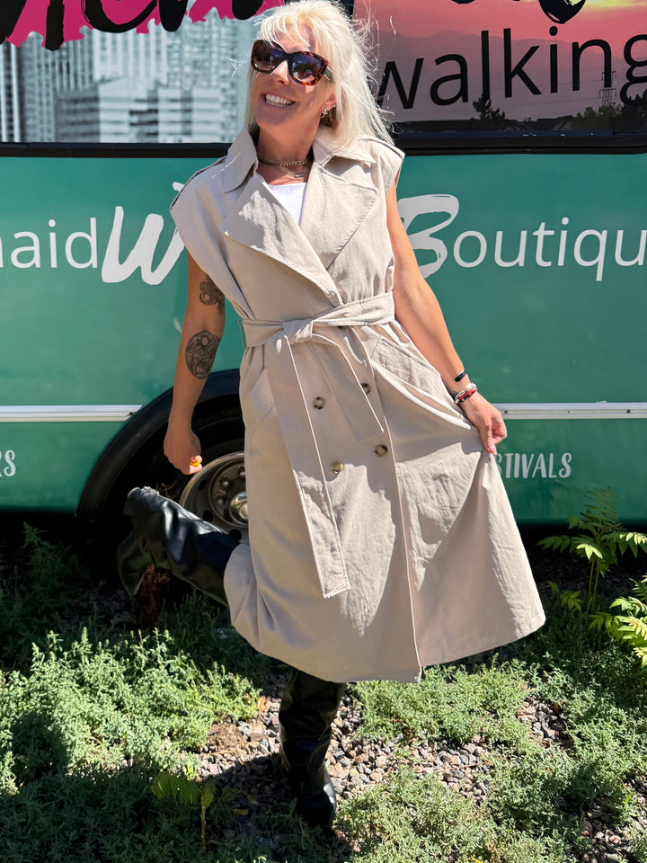 A woman wearing a beige trench coat and sunglasses stands in front of a teal bus with &