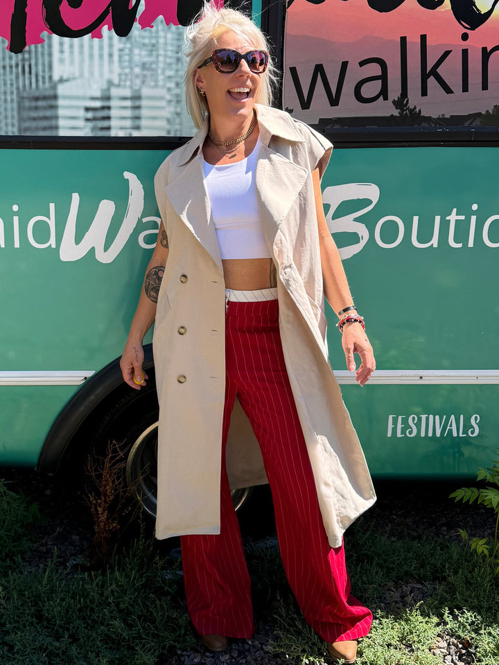 A woman stands outdoors in front of a teal bus with large windows and colorful Mermaid-themed graphics. She is wearing a beige trench coat over a white top and red wide-leg pants.