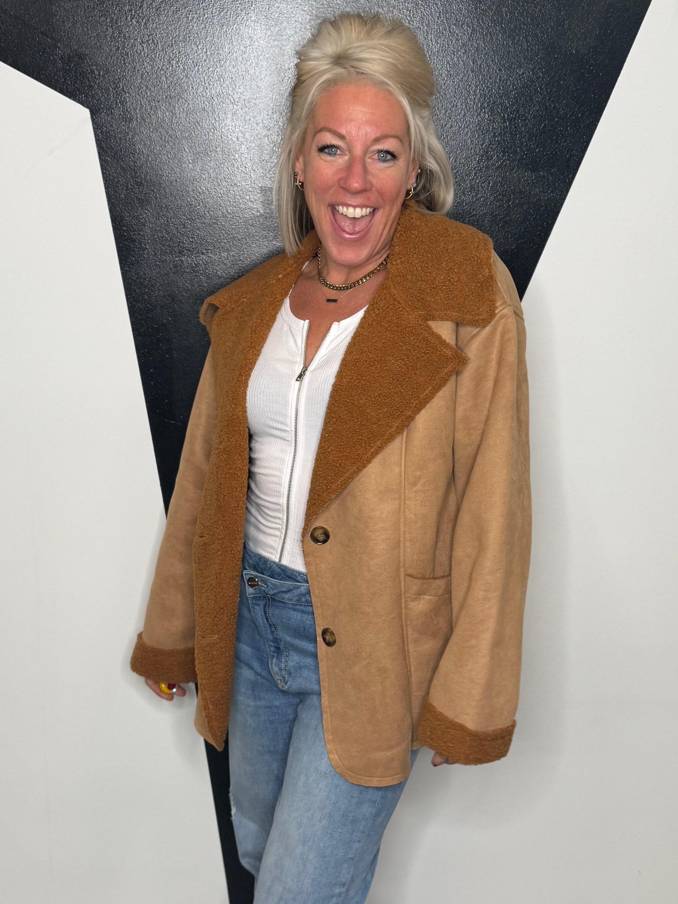 Woman wearing a brown coat and blue jeans standing against a black and white wall.