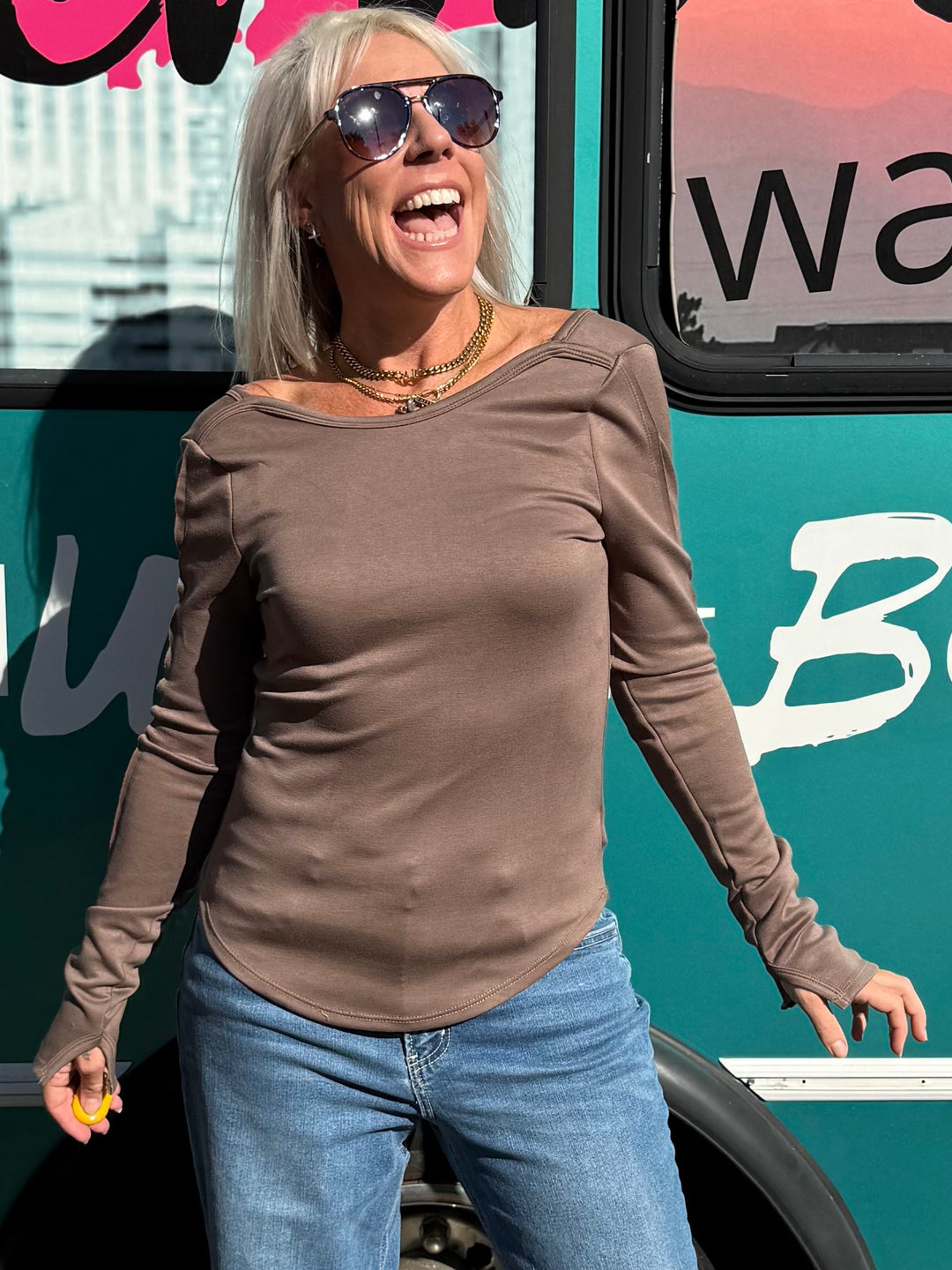 A woman wearing a brown long-sleeve top and blue jeans stands in front of a teal bus with &
