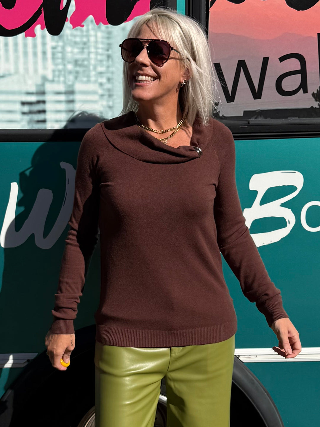 A woman wearing a brown cowl-neck long-sleeve top and olive green wide-leg pants stands in front of a teal bus with &