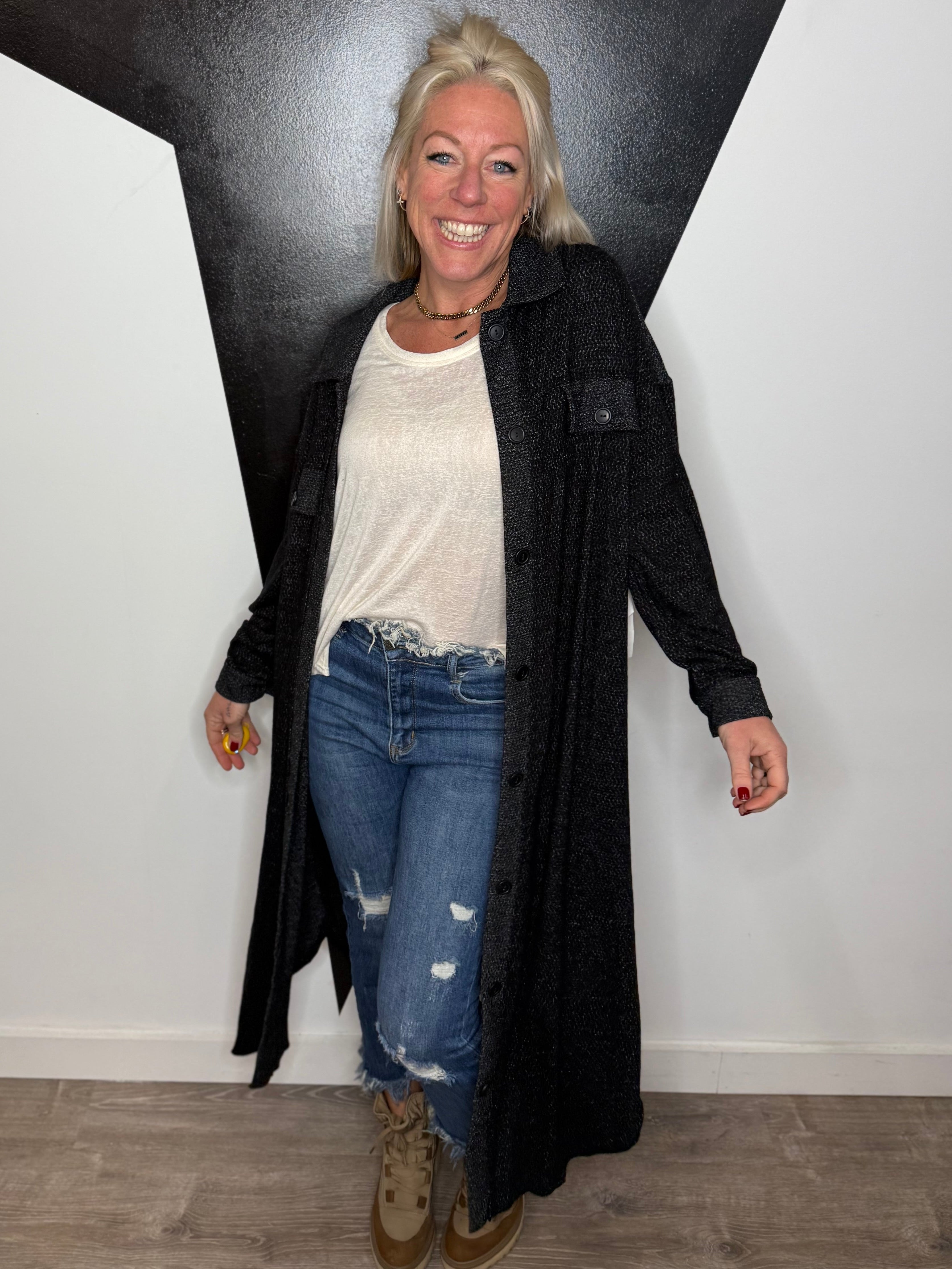 Person wearing a long black coat, white shirt, and blue jeans standing against a black and white wall.