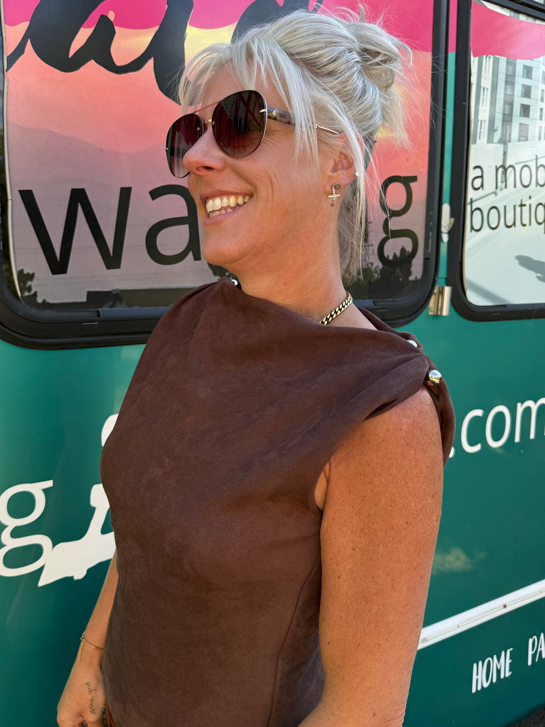 A smiling blonde woman wearing a brown sleeveless top and sunglasses stands in front of a green mobile boutique vehicle with pink and white graphics.