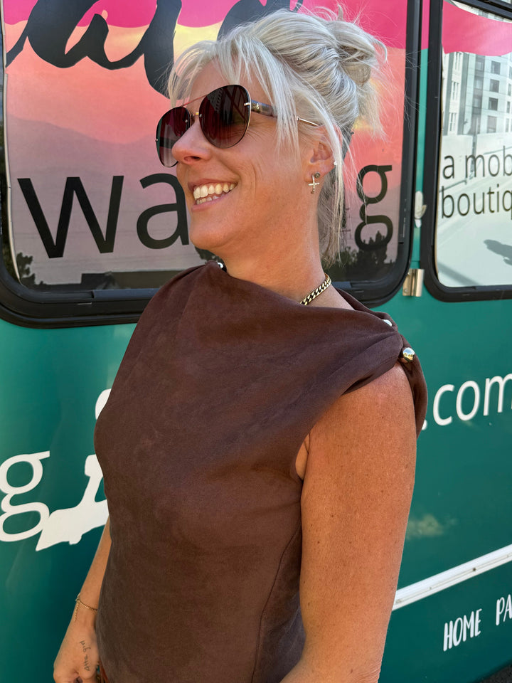 A smiling blonde woman wearing a brown sleeveless top and sunglasses stands in front of a green mobile boutique vehicle with pink and white graphics.