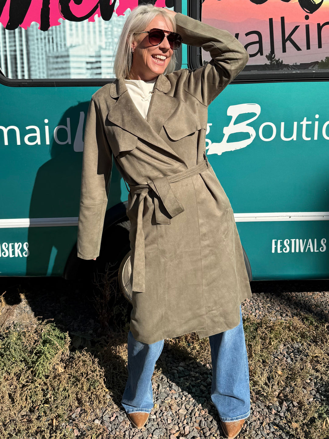 A person wearing a long olive freen trench coat, blue jeans, and sunglasses poses in front of a large teal and pink &