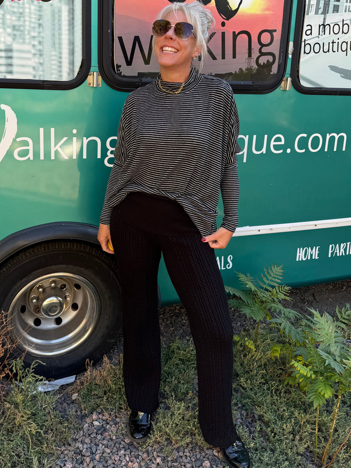 A woman wearing a black and white striped long-sleeve top and black pleated pants stands in front of a teal mobile boutique bus with pink and white text.
