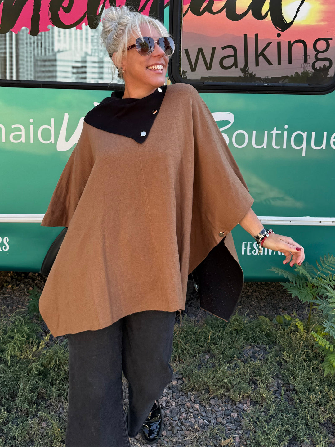 A woman wearing a tan poncho-style top with a black collar stands in front of a teal bus with &