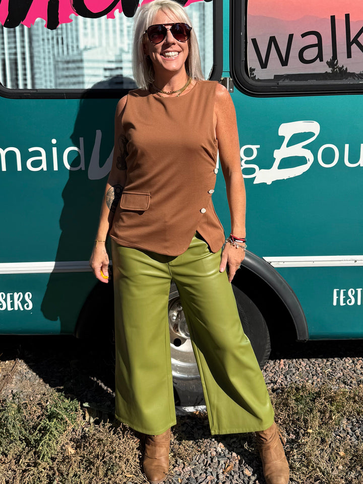 A woman wearing a brown asymmetrical top and olive green wide-leg pants stands in front of a teal bus with &