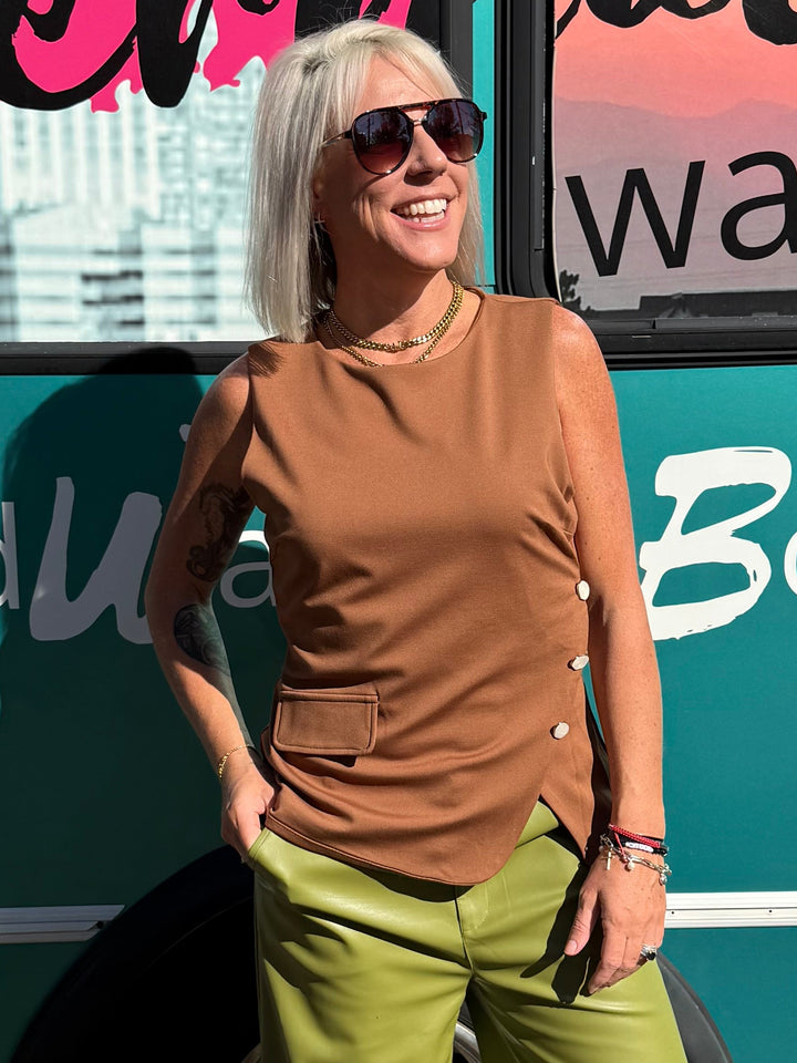 A woman stands outdoors in front of a teal bus with large windows and colorful window decals. She is wearing a sleeveless brown top, olive green wide-leg pants, and tan boots.