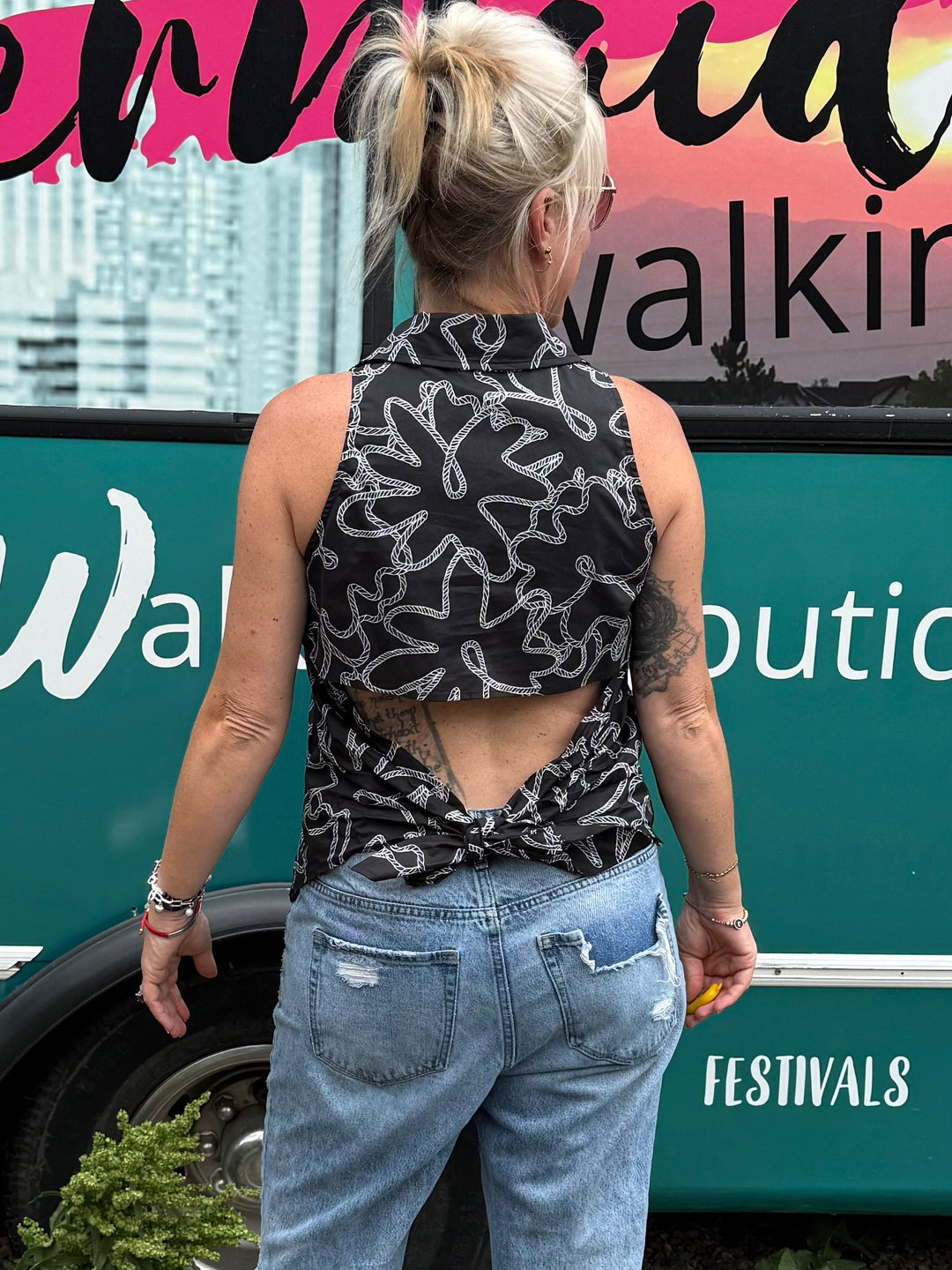 A woman stands with her back to the camera in front of a teal mobile boutique bus, wearing a black and white patterned halter top with a cutout back, light blue jeans, and black sandals. The bus has &