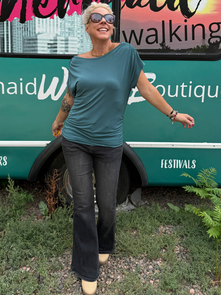 A woman wearing a teal short-sleeve t-shirt, dark flared jeans, and beige heels stands in front of a teal bus with &