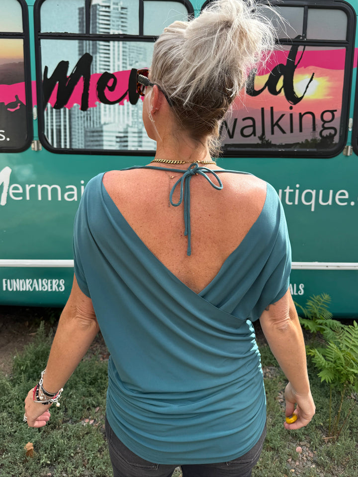A woman viewed from behind wearing a teal blue wrap-style top with a tie-back neckline, standing in front of a teal bus with &