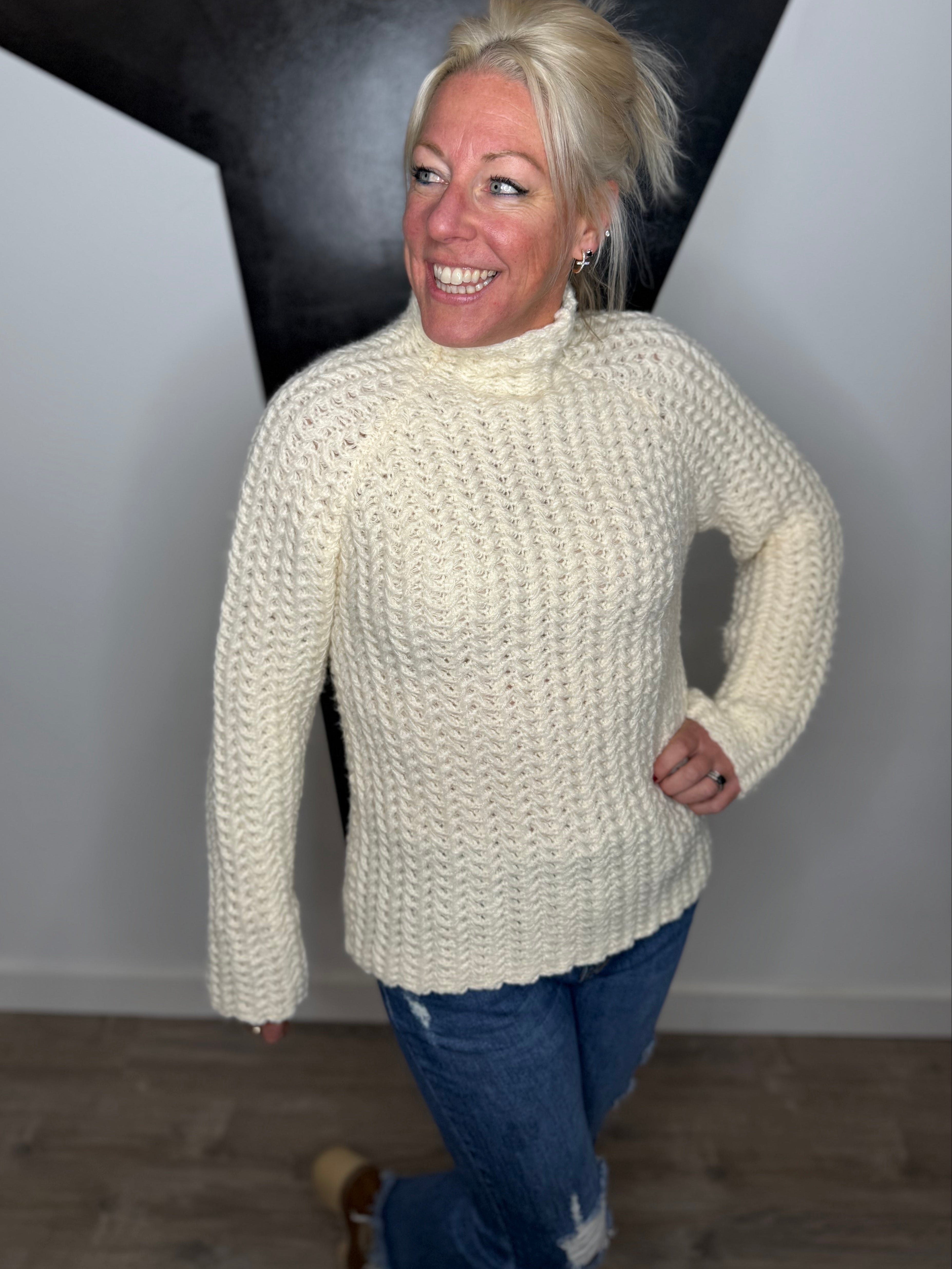 Woman wearing a white knitted sweater and blue jeans standing against a plain wall.