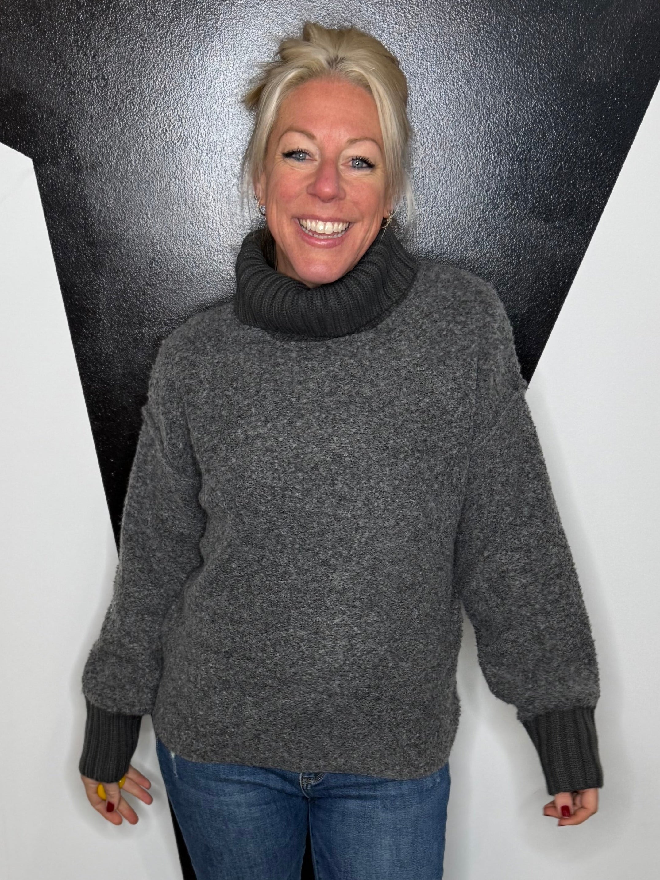 Person wearing a gray sweater and blue jeans standing against a black and white geometric wall.