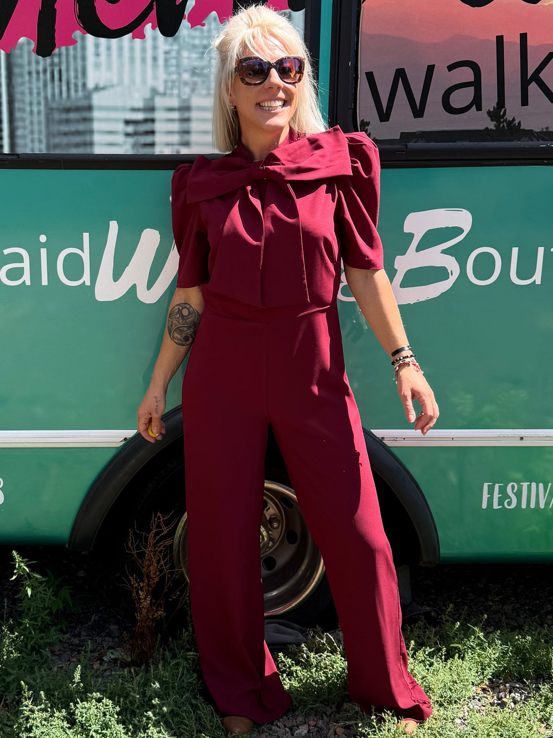 A woman wearing a burgundy jumpsuit and sunglasses stands in front of a teal bus with &