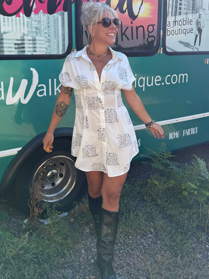 A woman wearing a white short-sleeve button-up shirt dress with black script print, black knee-high boots, and sunglasses, standing in front of a green mobile boutique truck.