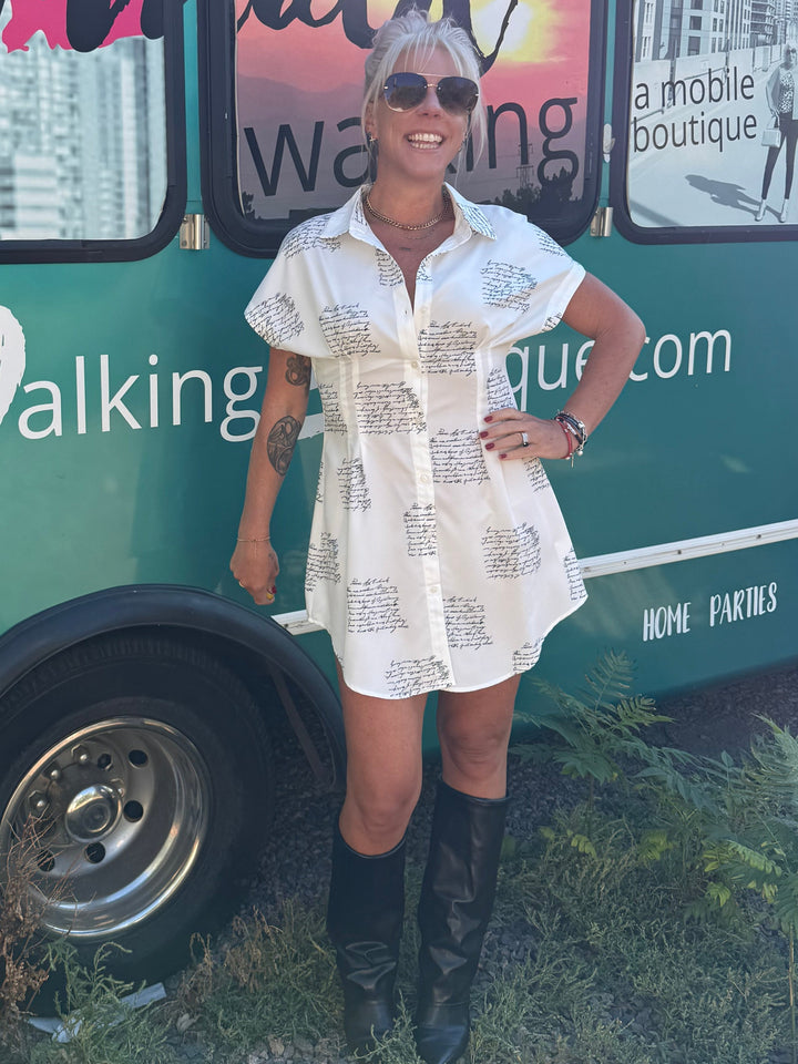 A woman wearing a white patterned short-sleeve shirt dress and black cowboy boots stands in front of a green bus with &