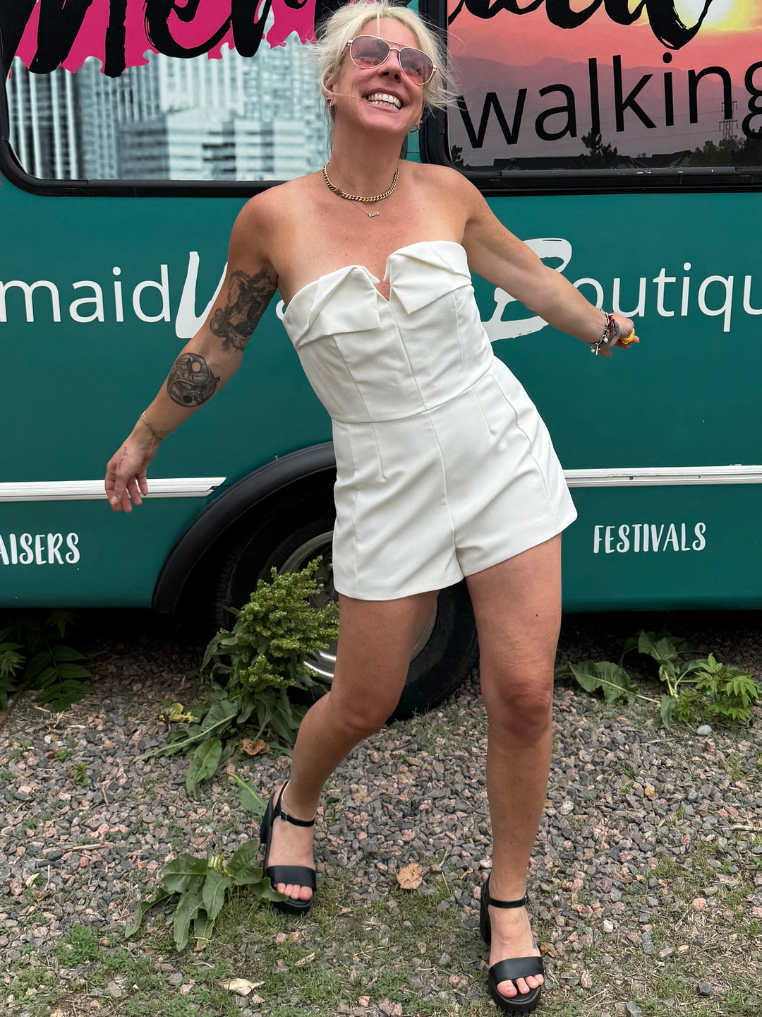 A woman posing outdoors in front of a teal bus with 'Mermaid Walking Boutique' written on the side. She is wearing a white strapless romper and black high-heeled sandals.